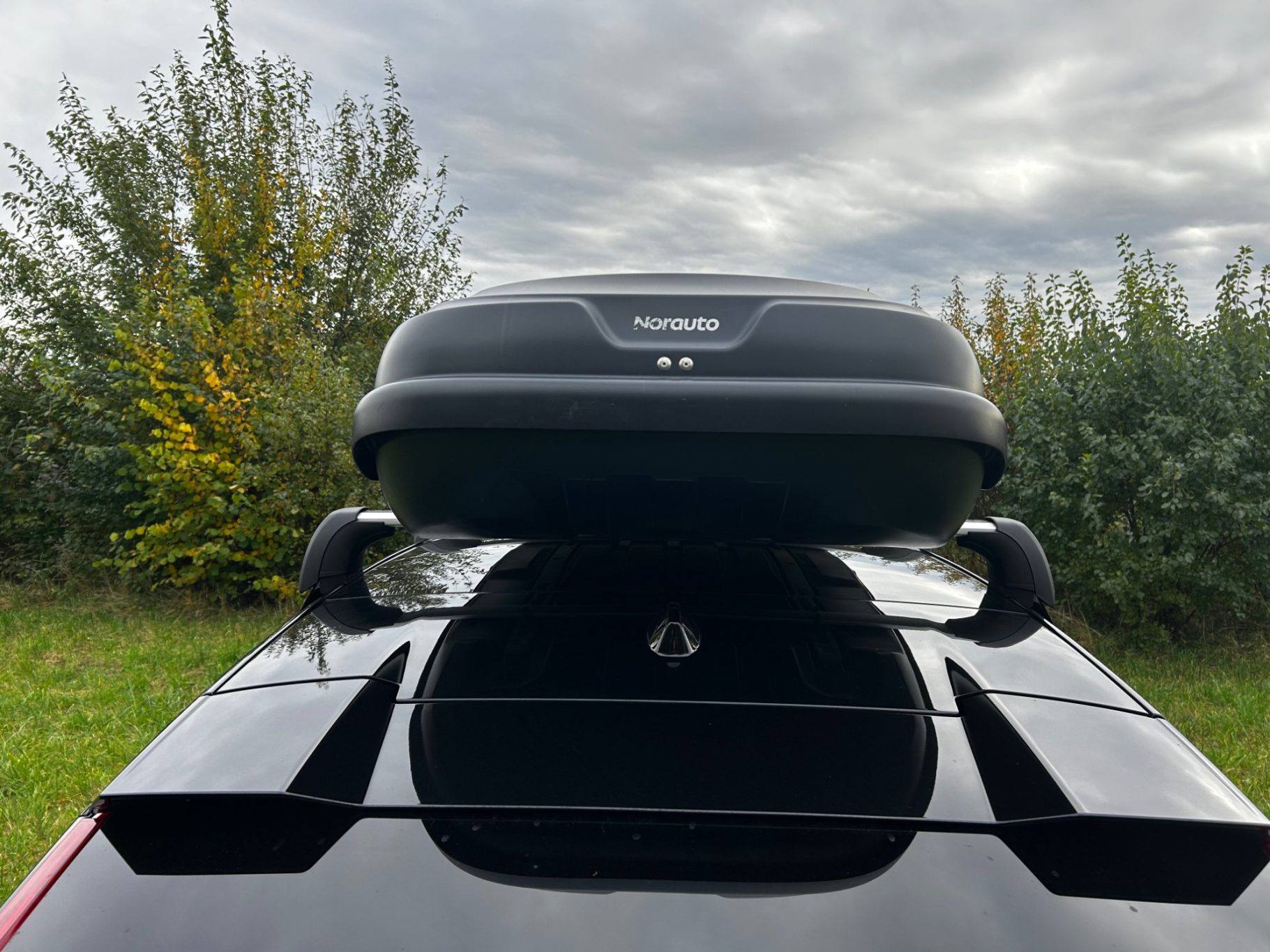 Dachbox von hinten Dachbox auf schwarzem Auto, im Hintergrund ein bewaldetes Gelände unter bewölktem Himmel.