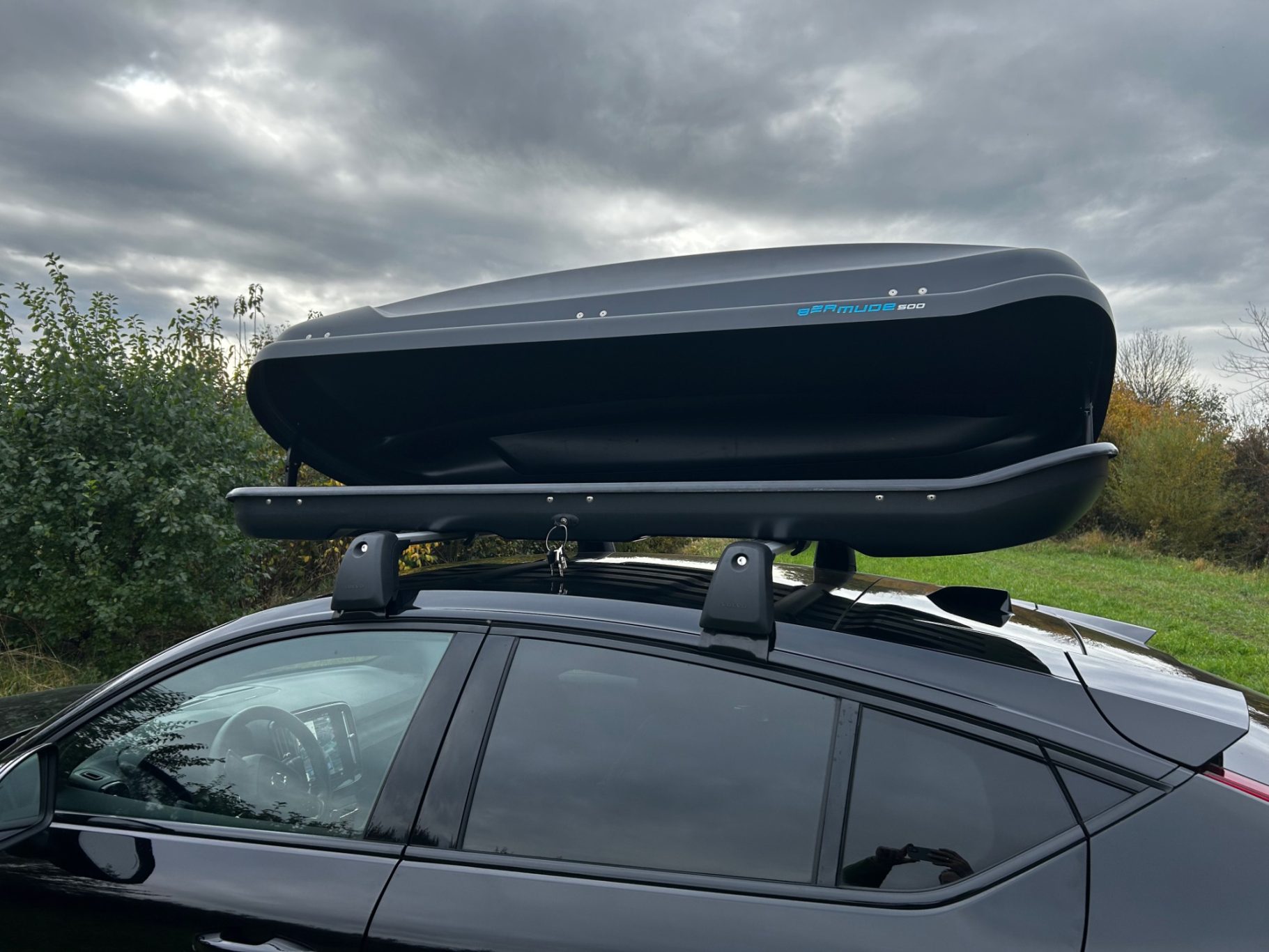 Dachbox geöffnet Fahrerseite Dachbox auf dem Gepäckträger eines Autos, vor bewölktem Himmel und Bäumen.
