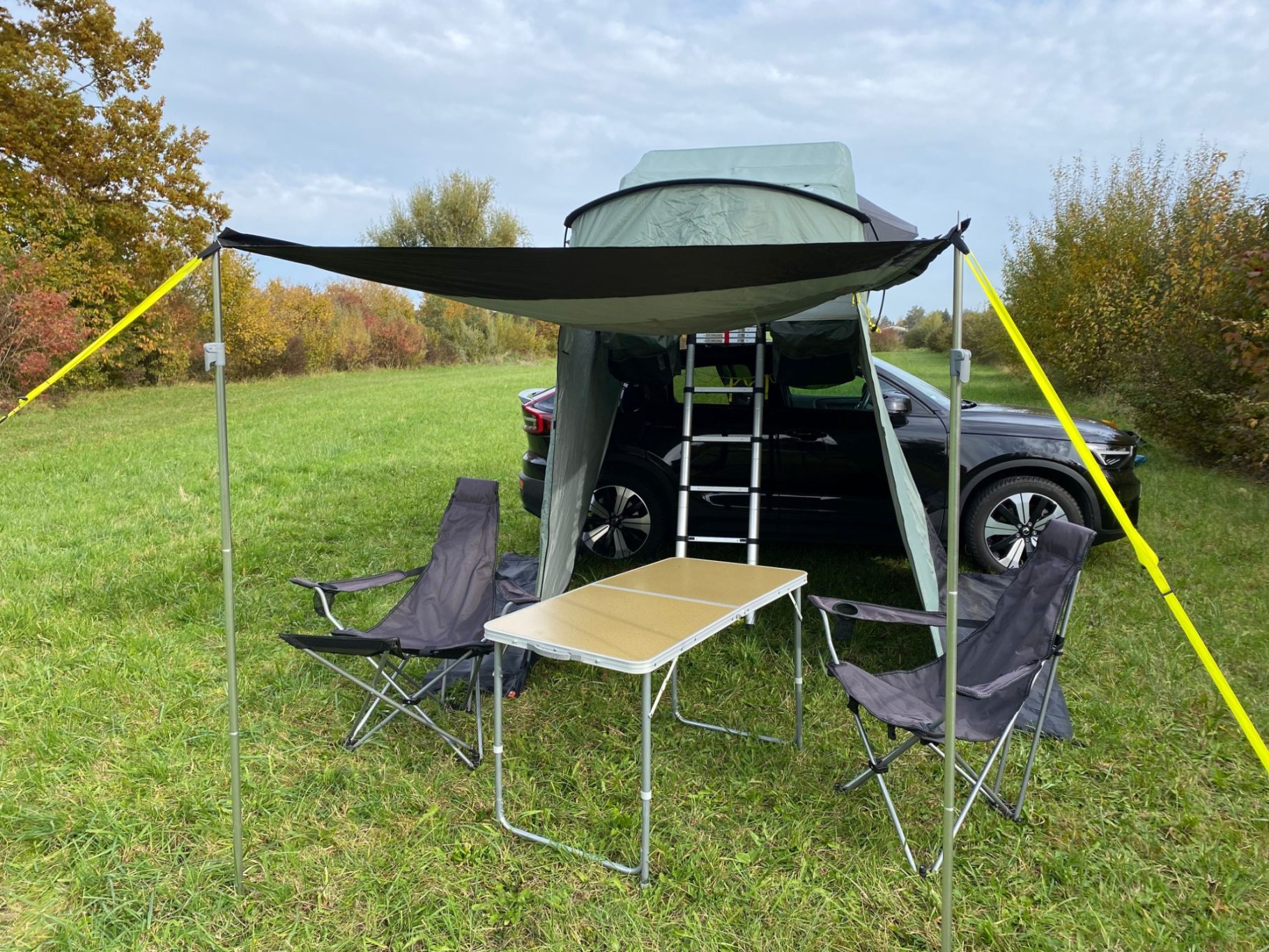 Vorzelt als Markise geöffnet Zelt mit Vordach, zwei Campingstühlen und einem Tisch auf einer Wiese neben einem Auto.