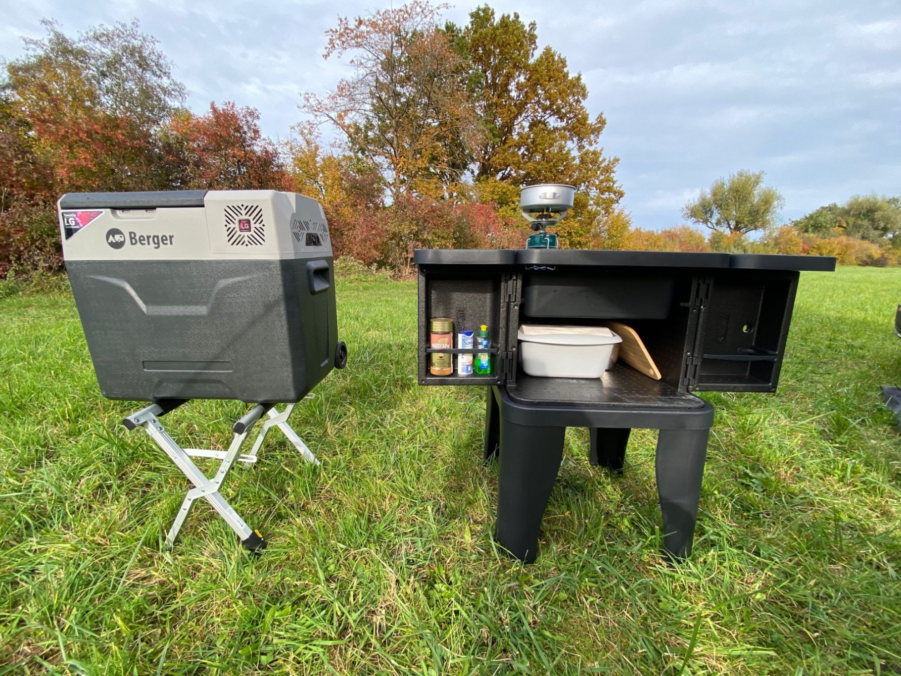 neben der Outdoorküche Kühlbox und Tisch stehen in einer grünen Wiese mit Bäumen im Hintergrund.
