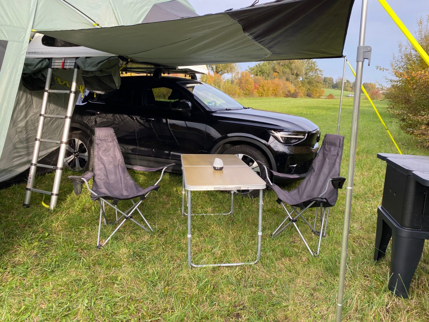 Vorzelt seitlich als Markise geöffnet Schwarzes Auto neben einem Campingzelt, mit zwei Stühlen und einem Tisch im Freien.