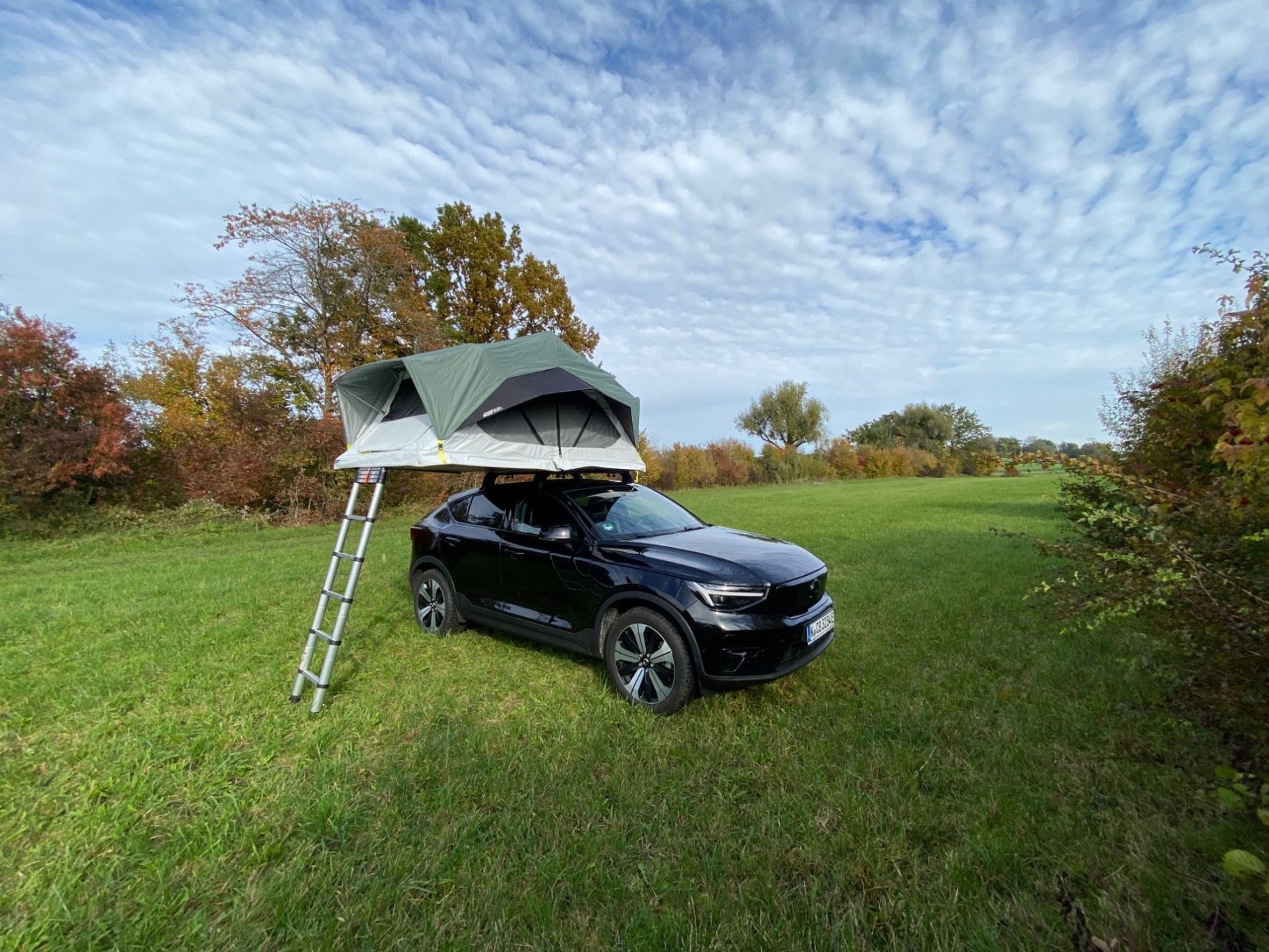 Dachzelt MH500 Schwarzes Auto mit aufklappbarem Dachzelt in einer grünen Wiese unter einem bewölkten Himmel.