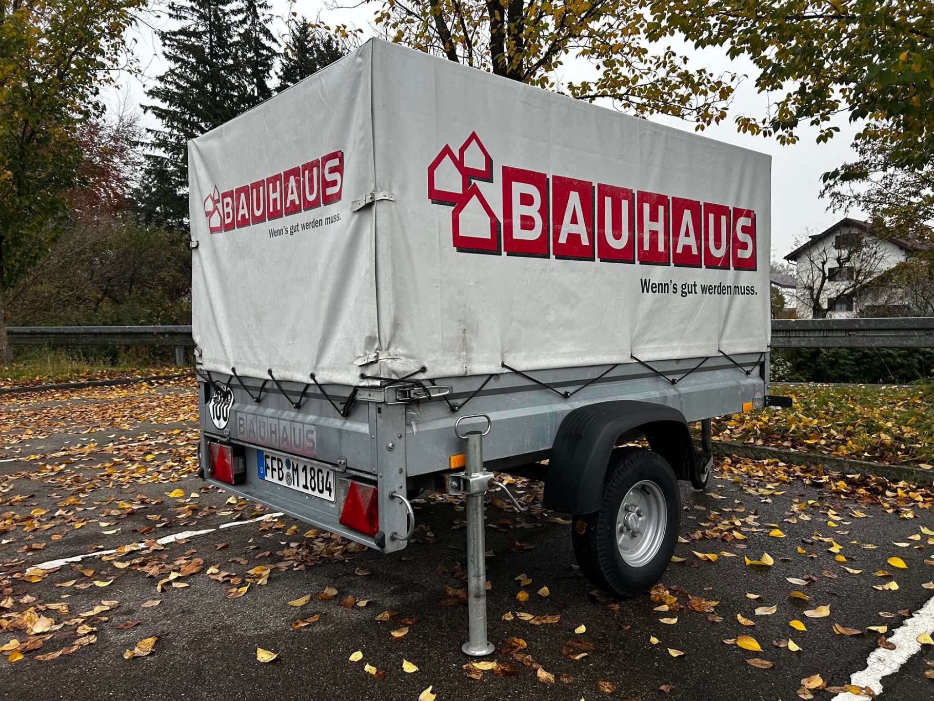 Anhänger mit Stütze PKW-Anhänger mit Bauhaus-Logo, abgedeckt, in einem herbstlichen Parkbereich.