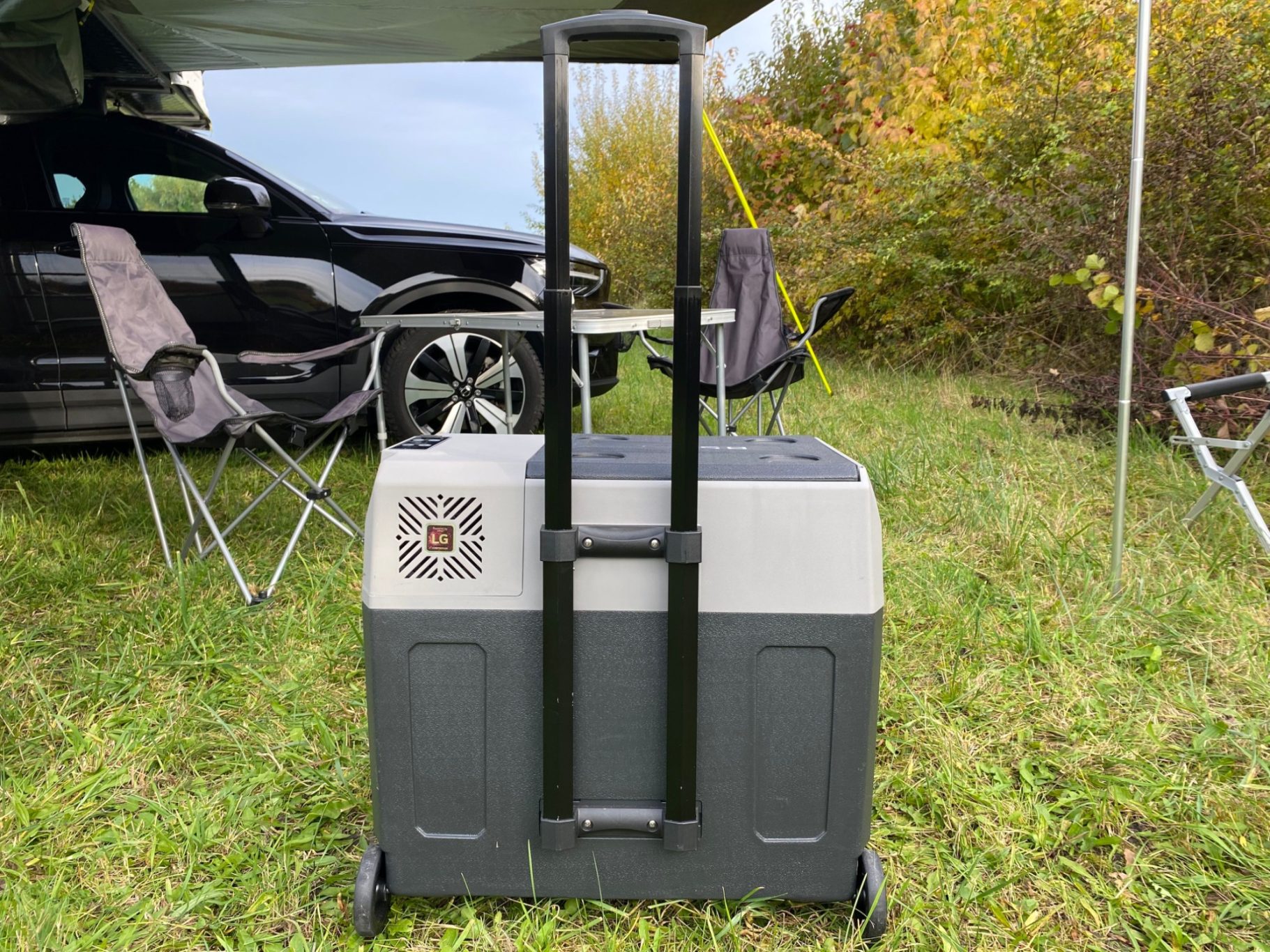 Trolleyfunktion zum leichten Transport Kühlbox mit Rollen und Griff, im Freien neben Campingstühlen und einem Auto.