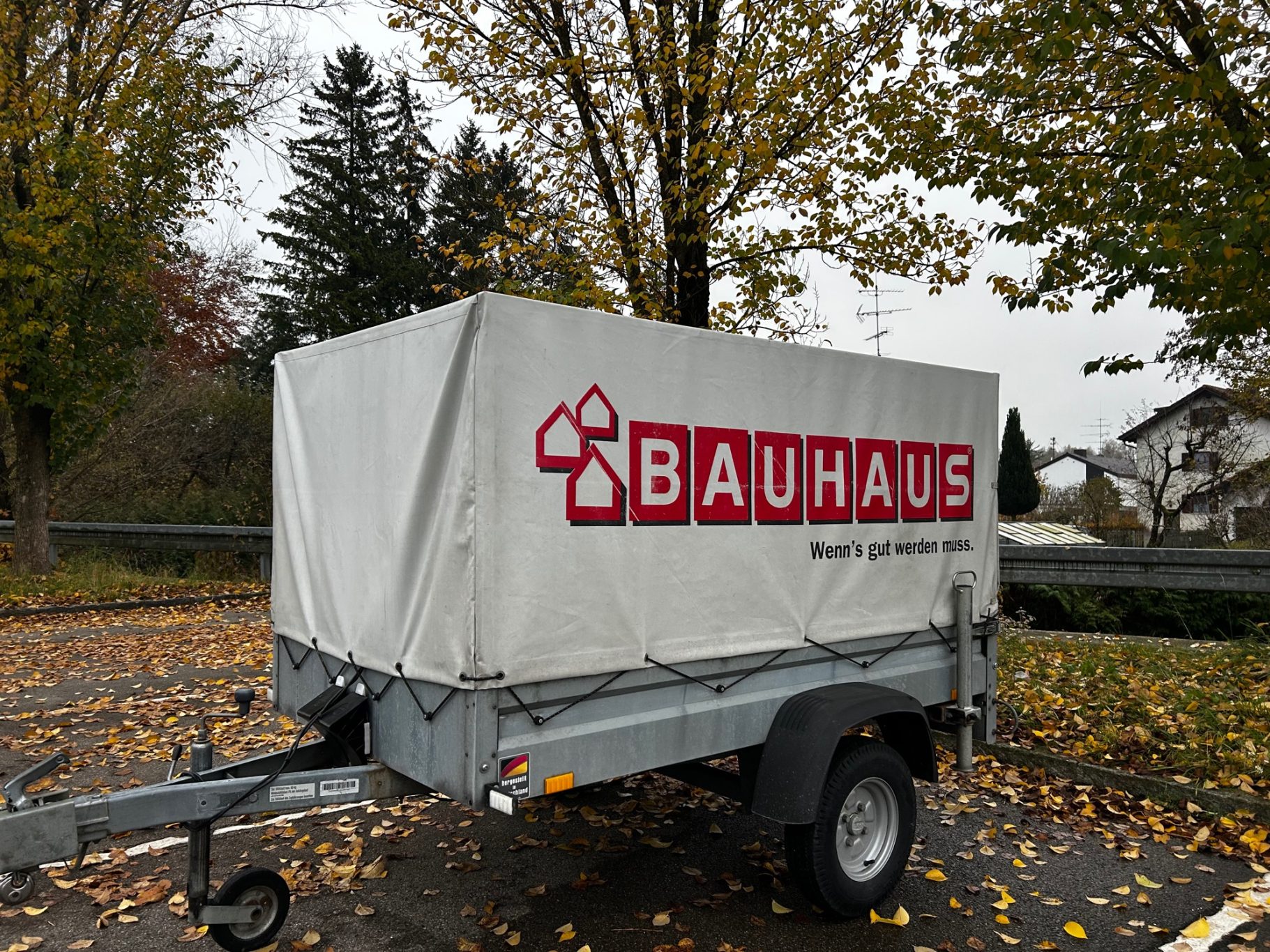 PKW-Anhänger vorne Bauhaus-Anhänger mit grauem Planenaufbau und herbstlichem Laub im Hintergrund.