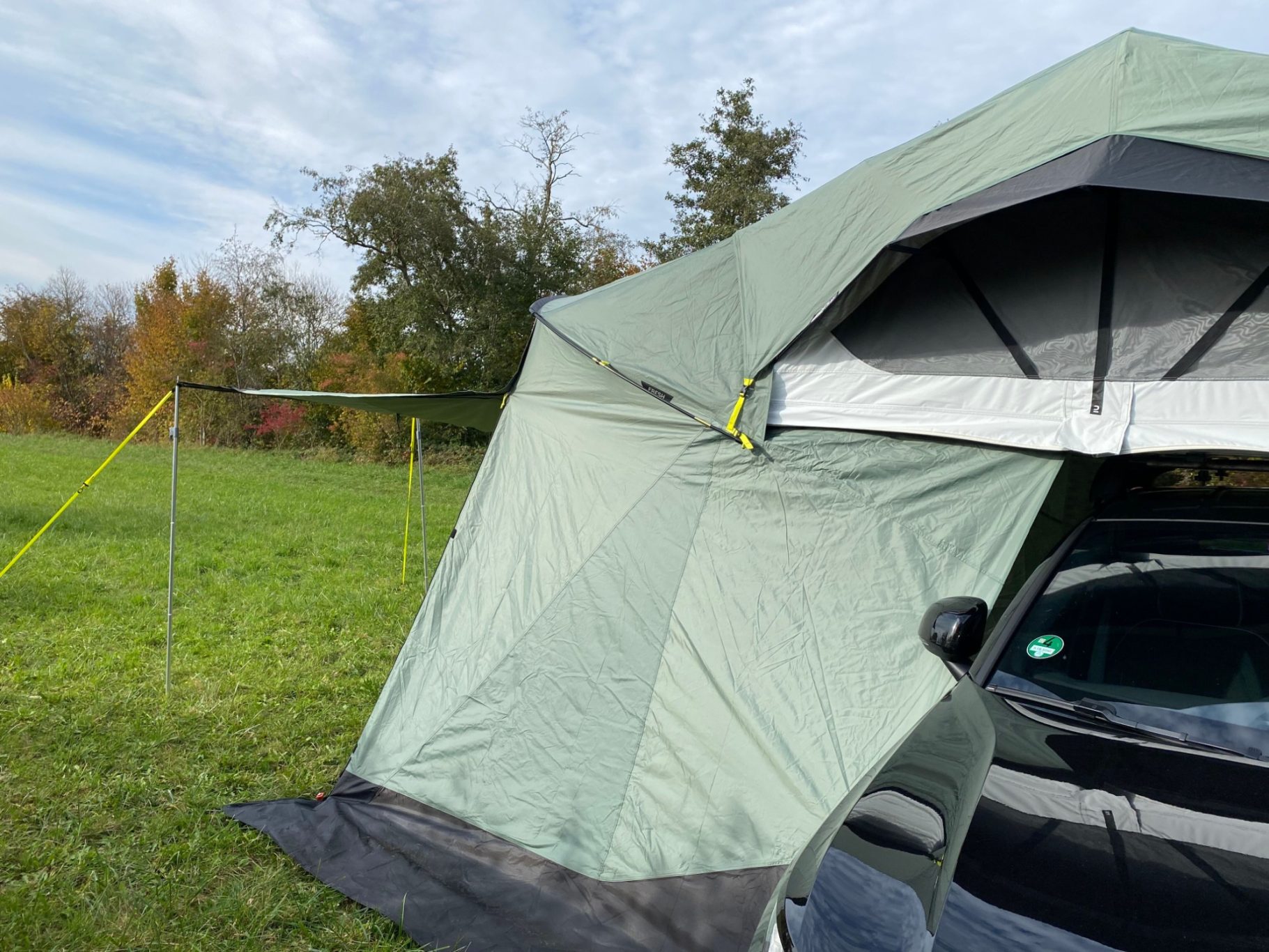 Vorzelt frontal als Markise geöffnet Zelt in einer Wiese mit einem Auto daneben und bewölktem Himmel im Hintergrund.
