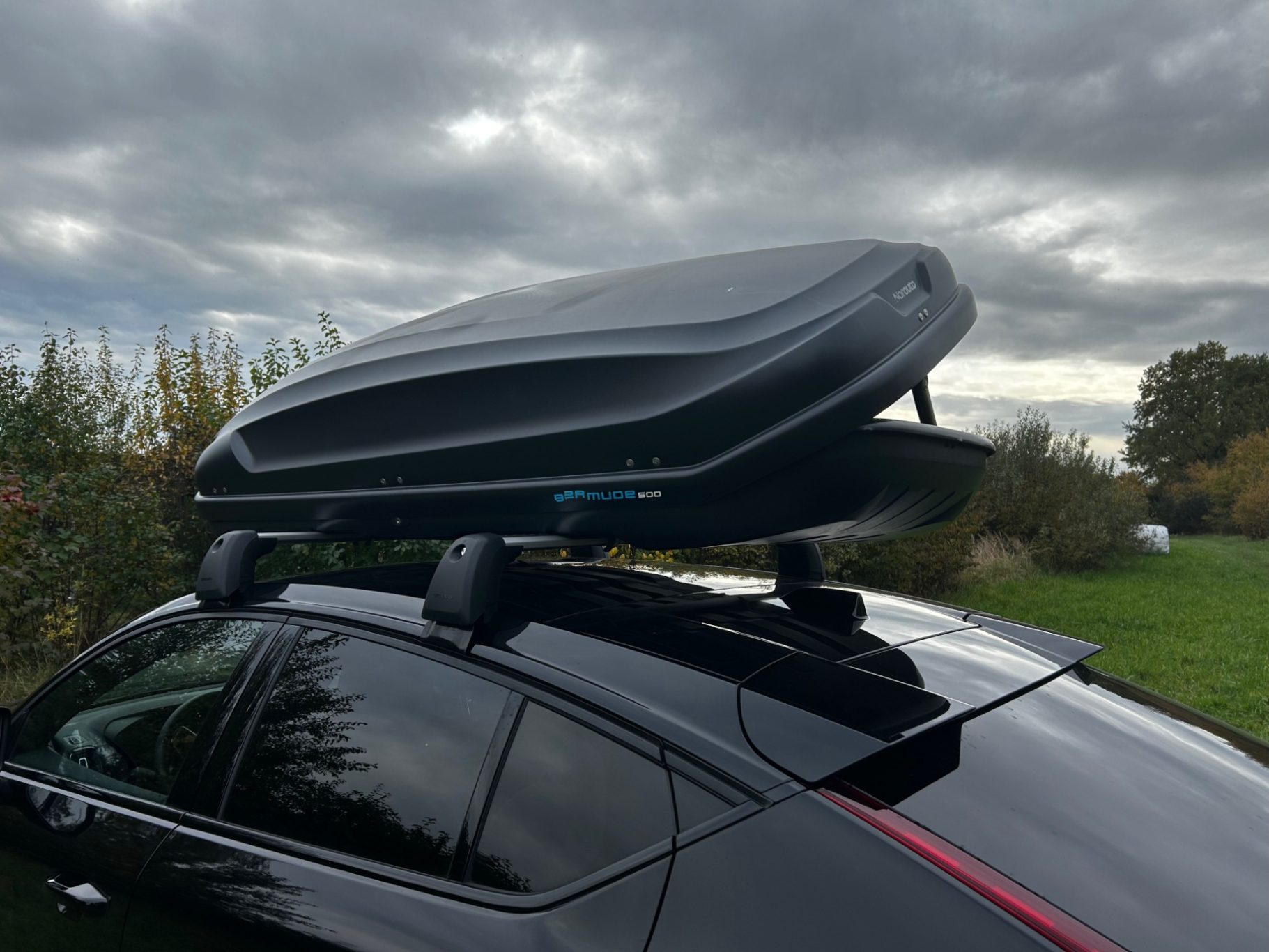 Dachbox geöffnet Beifahrerseite Dachbox auf dem Fahrzeugdach, über einem dunklen Auto, vor bewölktem Himmel.