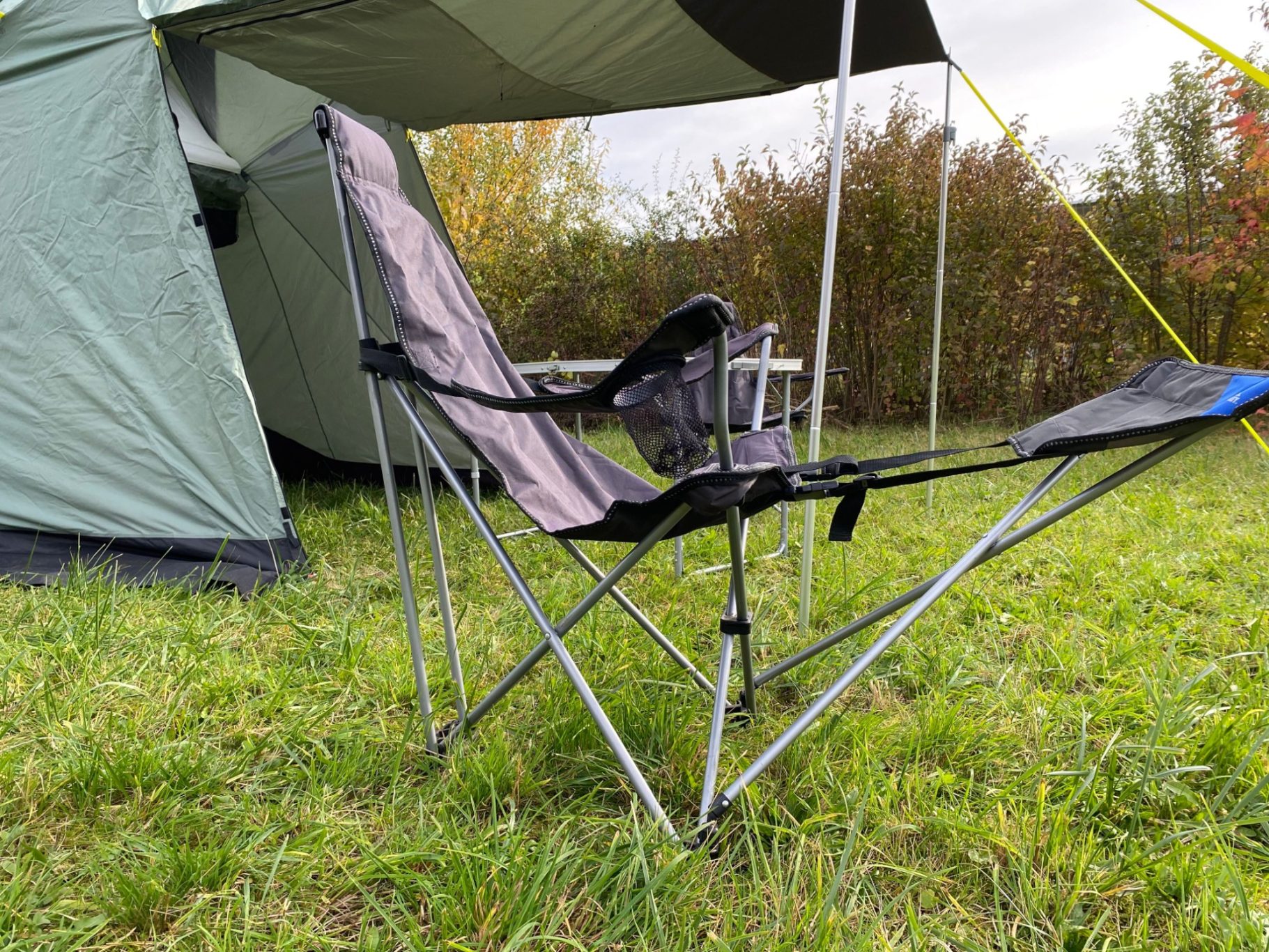McKinley Klappstuhl mit Fußteil Faltstuhl unter einem Zelt, umgeben von grünem Gras und Sträuchern.
