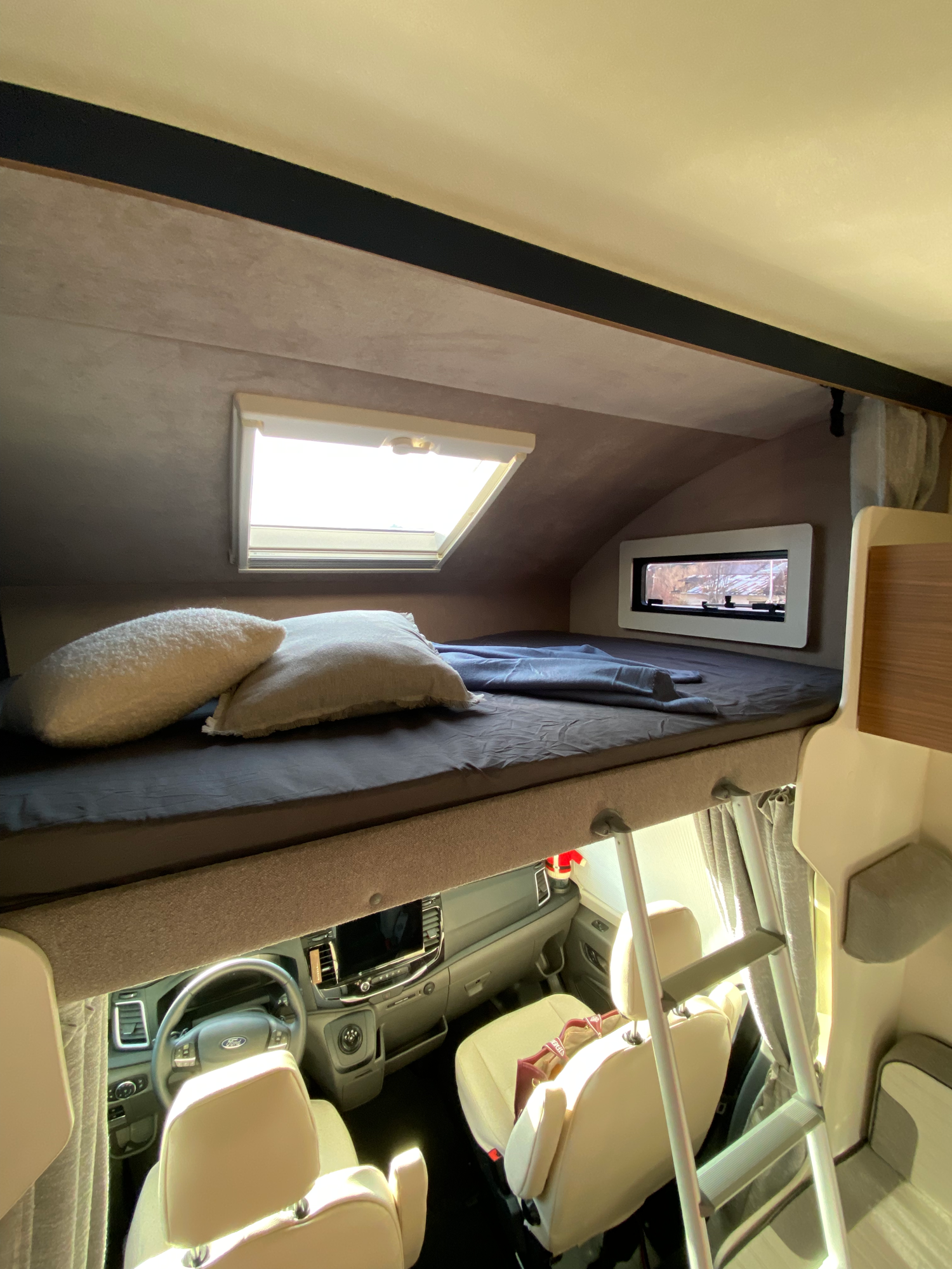 Alkoven mit zu öffnenden & verdunkelbaren Fenstern Hochbett in einem Wohnmobil mit Fenstern und Kissen, darunter Fahrersitz.
