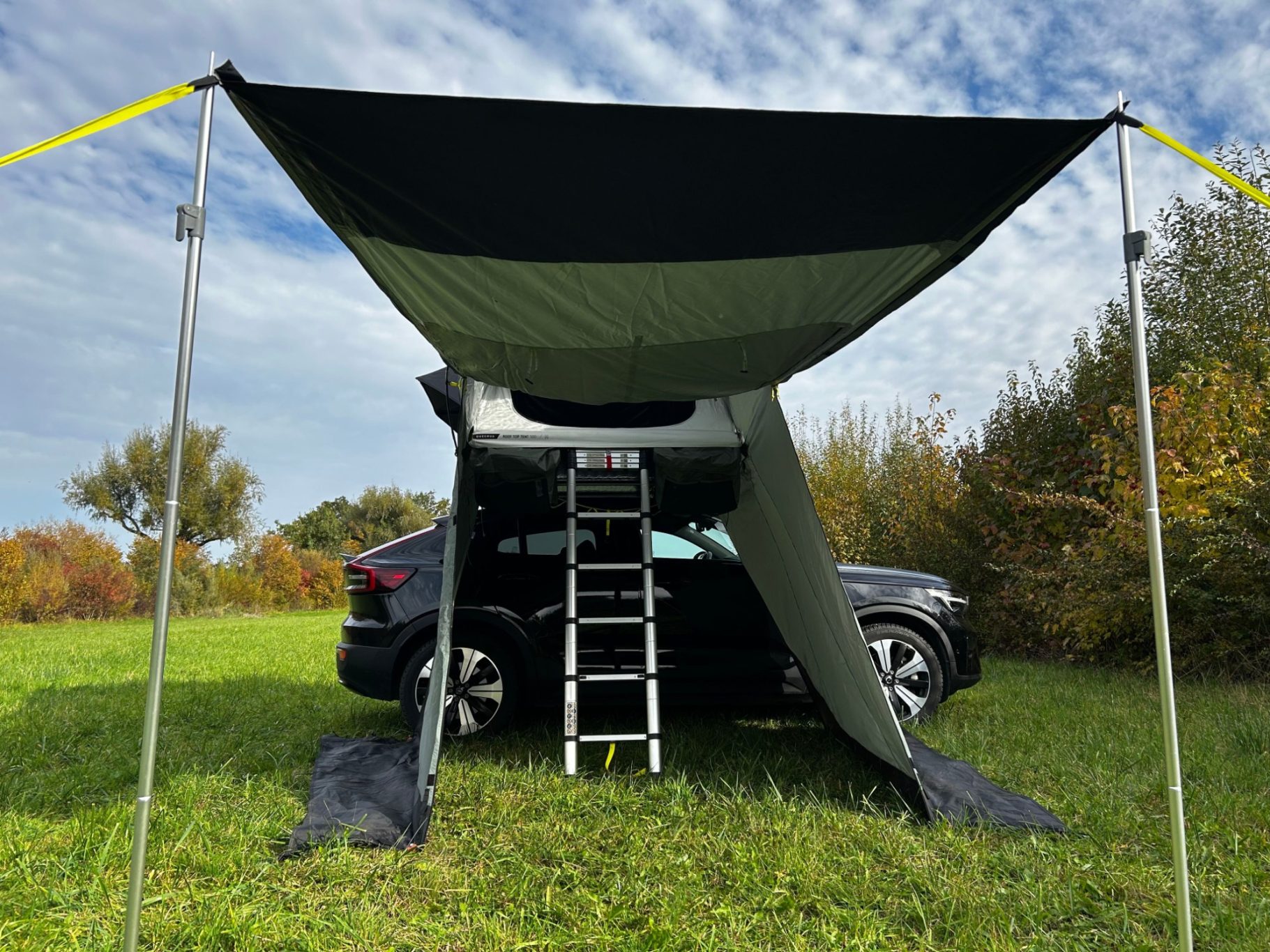 Vorzelt frontal als Markise geöffnet Zeltüberdachung über einem schwarzen Auto, mit einer Leiter und Grün im Hintergrund.