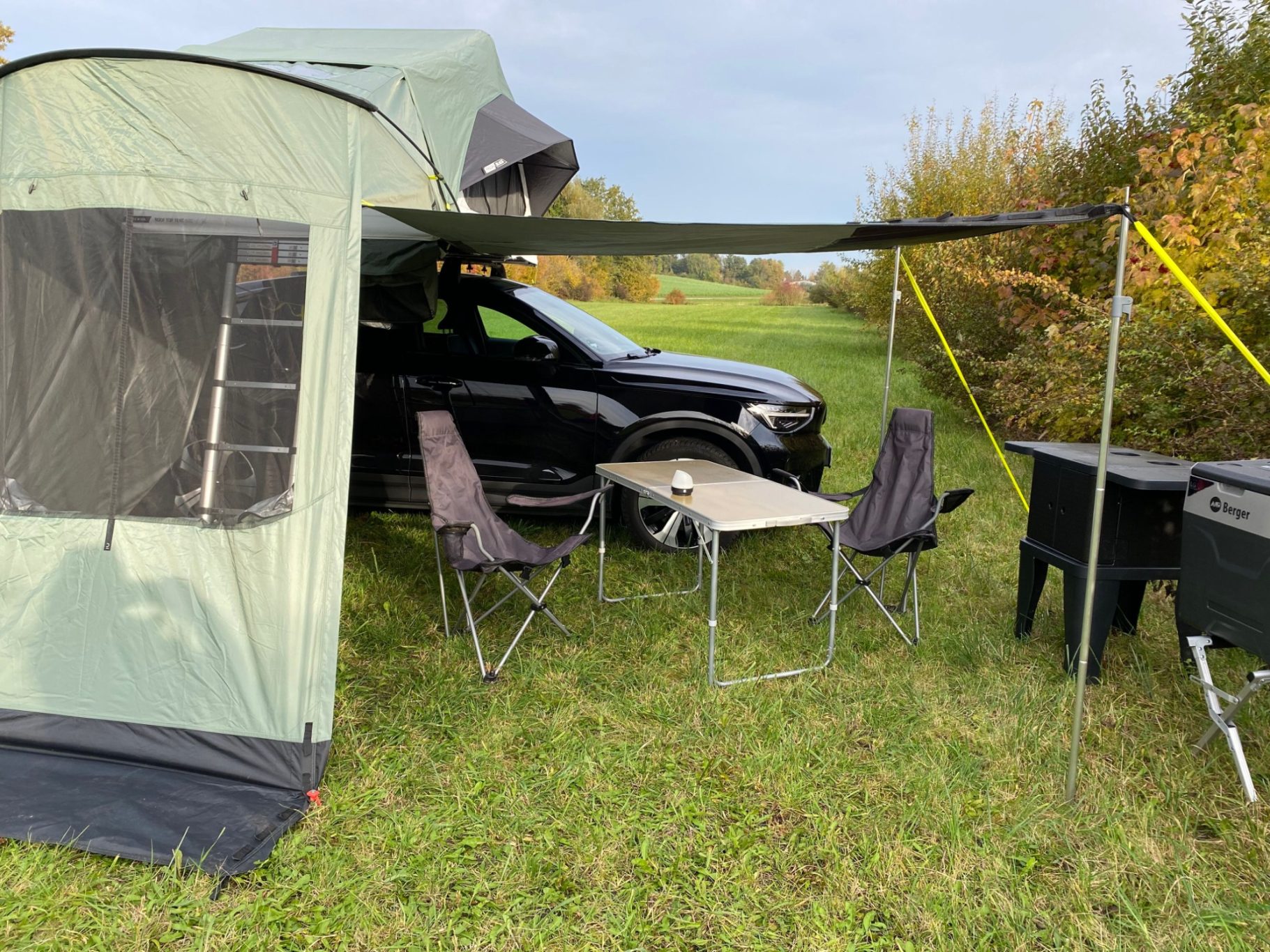 optionales Vorzelt geöffnet Zelt mit einem Fahrzeug, zwei Stühlen und einem Tisch auf einer Wiese in der Nähe von Bäumen.