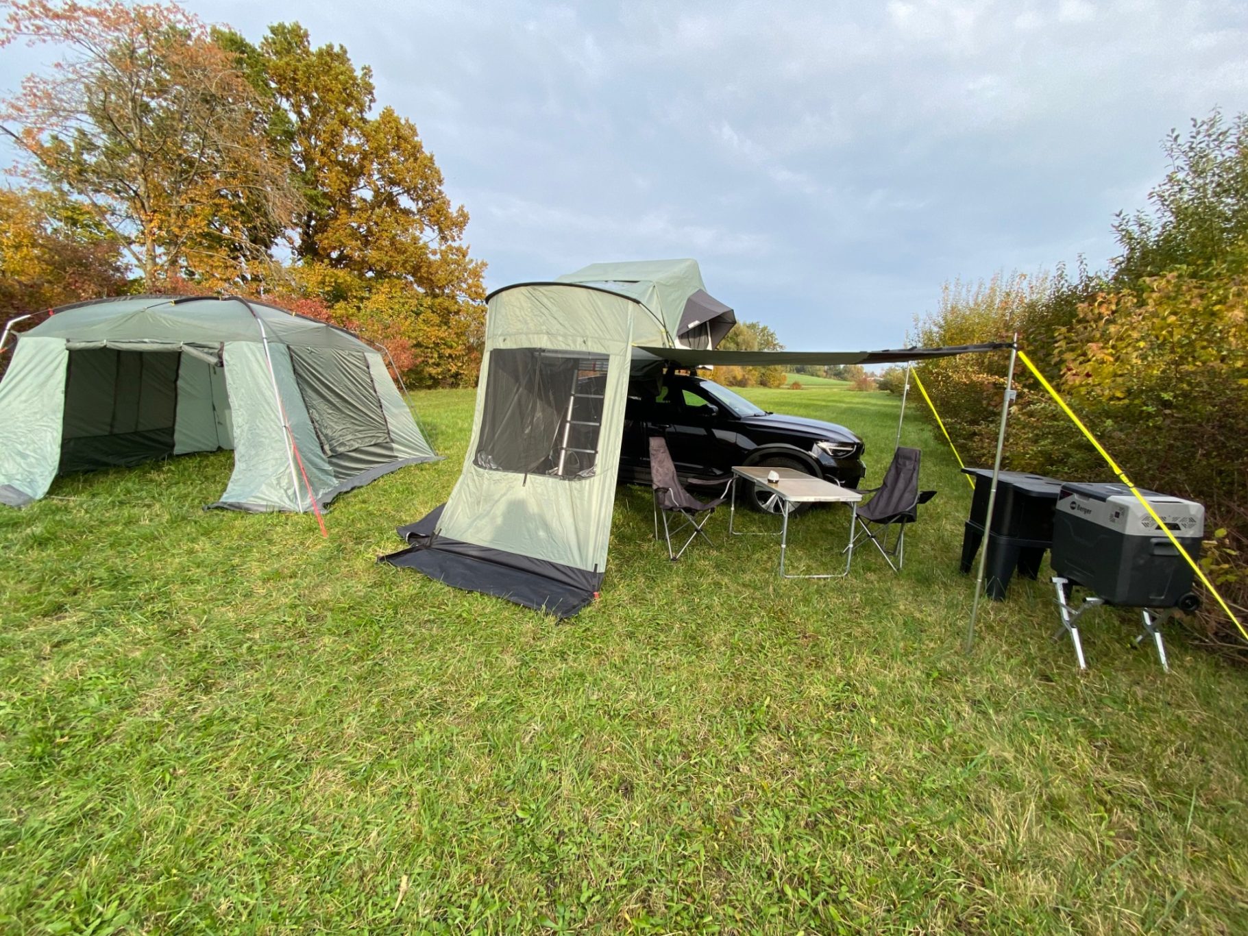 Perfekte Erweiterung Zwei Zelte auf einer Wiese, daneben ein Auto und Campingmöbel. Herbstliche Baumkulisse.